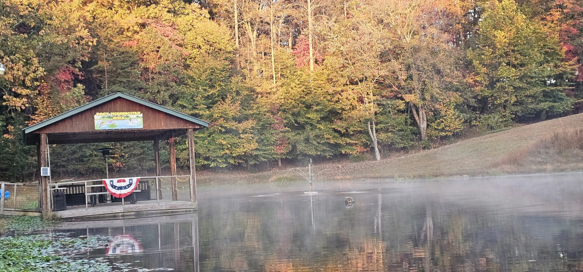 A misty lake at sunrise with autumn trees and a wooden dock.
