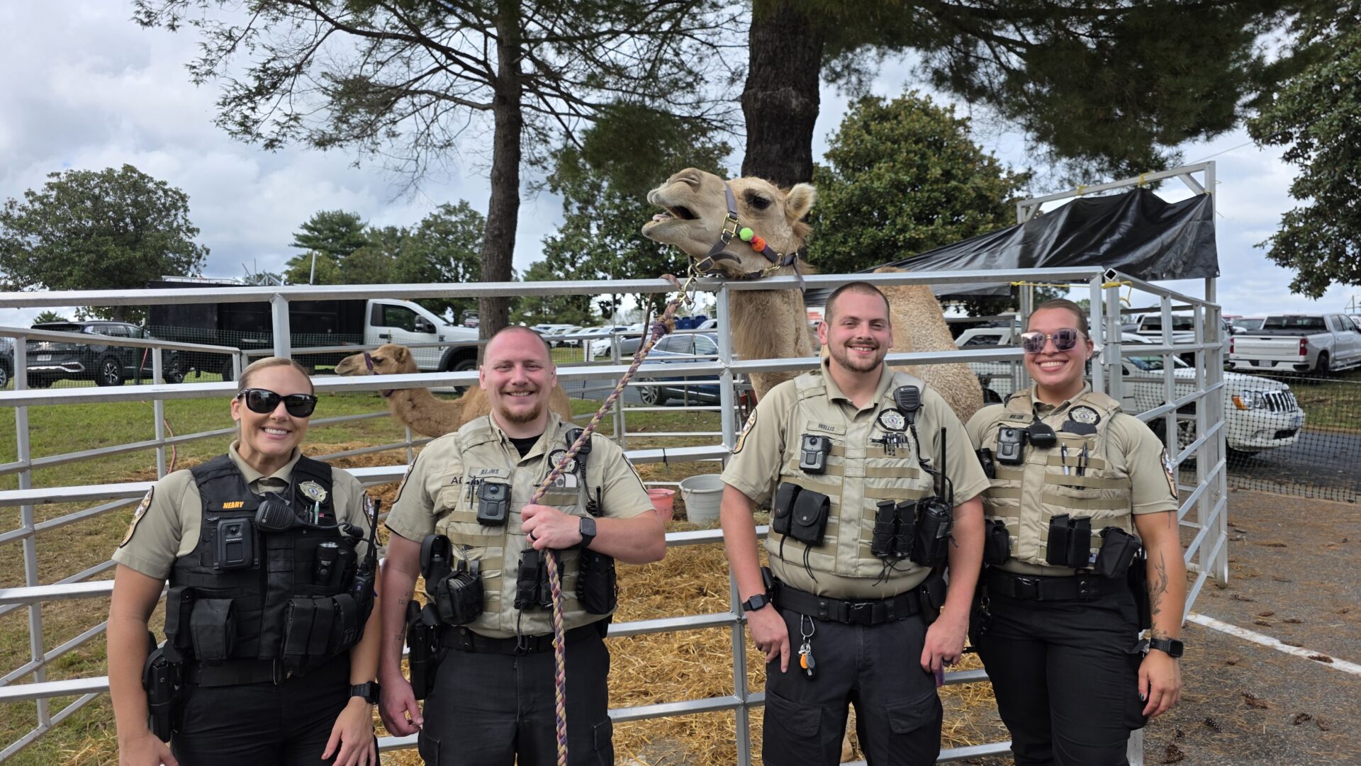 Three police officers smiling outdoors by a fence with a camel behind them.