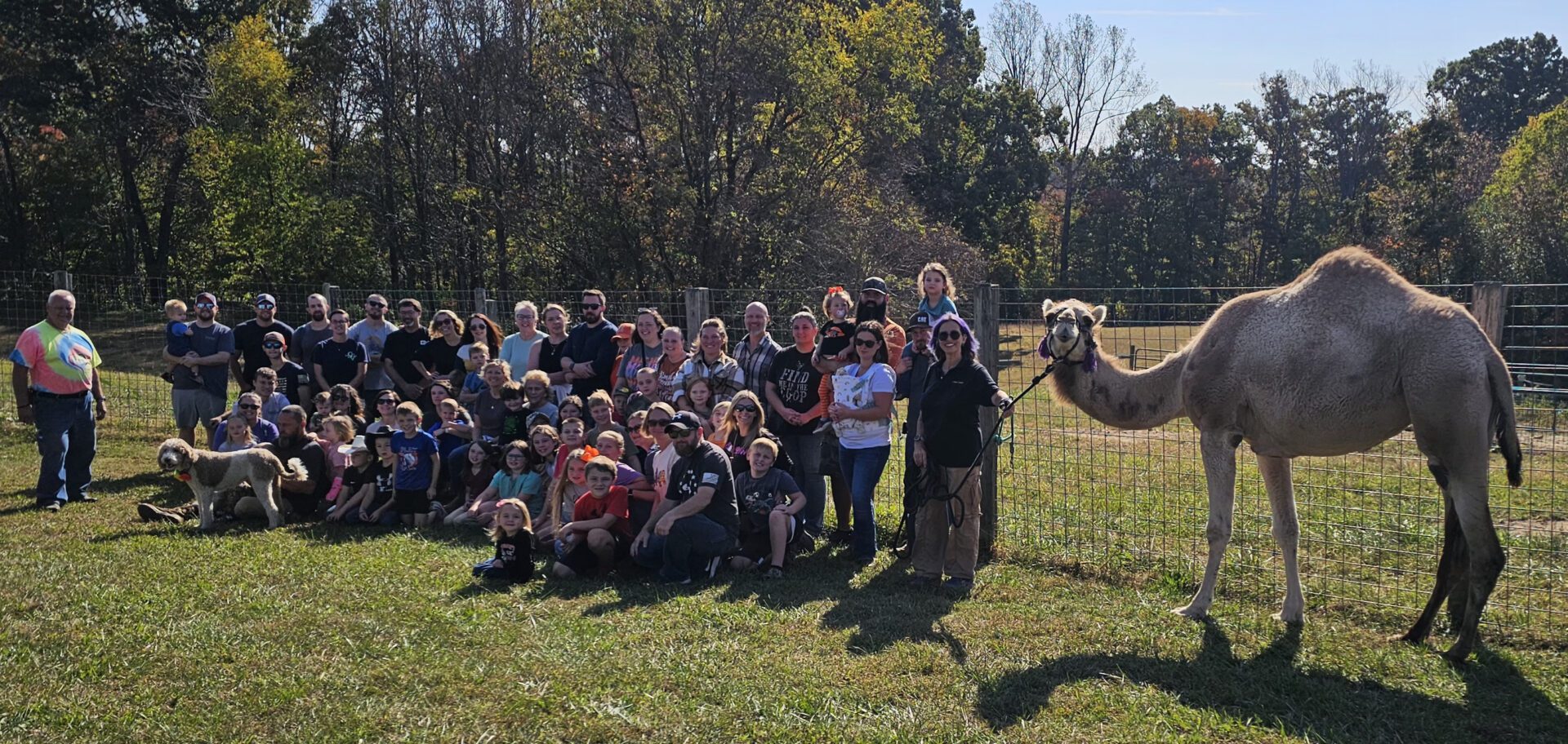 A large group of people posing outdoors with a llama during the day.