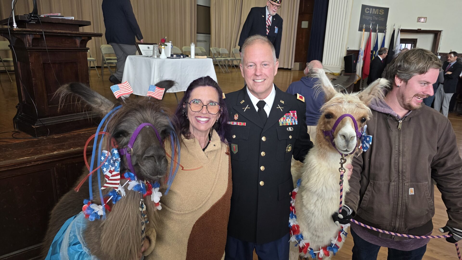 Man in military uniform and woman stand smiling with decorated llamas.